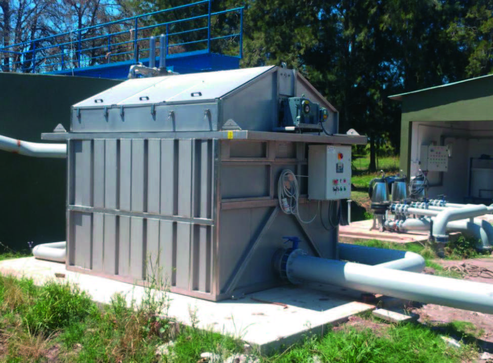 A large, rectangular industrial machine with metal panels and pipes is installed outdoors on a cement platform, surrounded by grass and trees. The unit features control panels and is connected to several large white pipes.