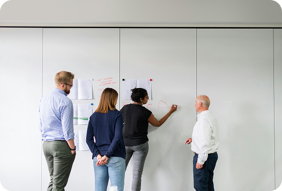 Four people stand in front of a whiteboard with papers and diagrams pinned to it. One person is writing or drawing on the board while the others watch and discuss. The scene appears to be a collaborative meeting or brainstorming session.
