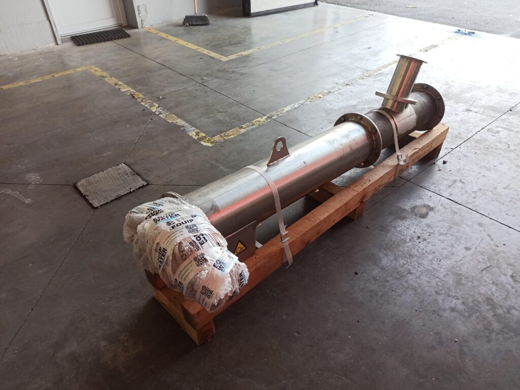 A large metal industrial pipe wrapped at one end sits secured on a wooden pallet in a warehouse with concrete floors and yellow safety markings.