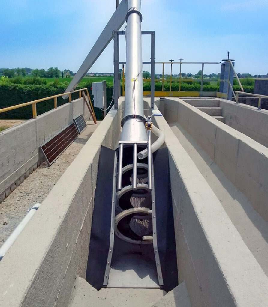 A large metal screw conveyor is installed within a concrete channel outdoors, surrounded by industrial structures and green fields under a clear blue sky.