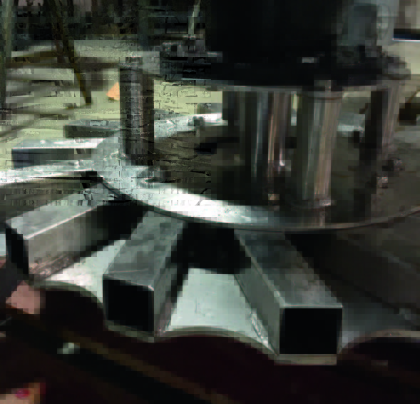 A close-up of a metal machine part with a circular base, vertical cylindrical supports, and rectangular metal spokes arranged radially. The image is taken in an industrial setting with dim lighting.