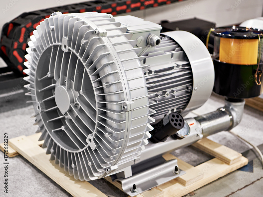 A close-up of an industrial electric air blower pump with a large metal housing and cooling fins, placed on a wooden base in a workshop or factory setting.