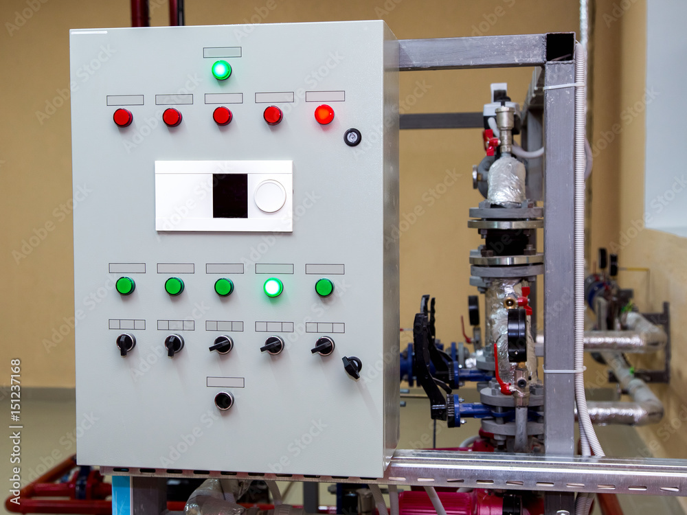 A control panel with various green and red indicator lights, switches, and a small display screen is mounted next to industrial pipes and valves in a mechanical or engineering setting.