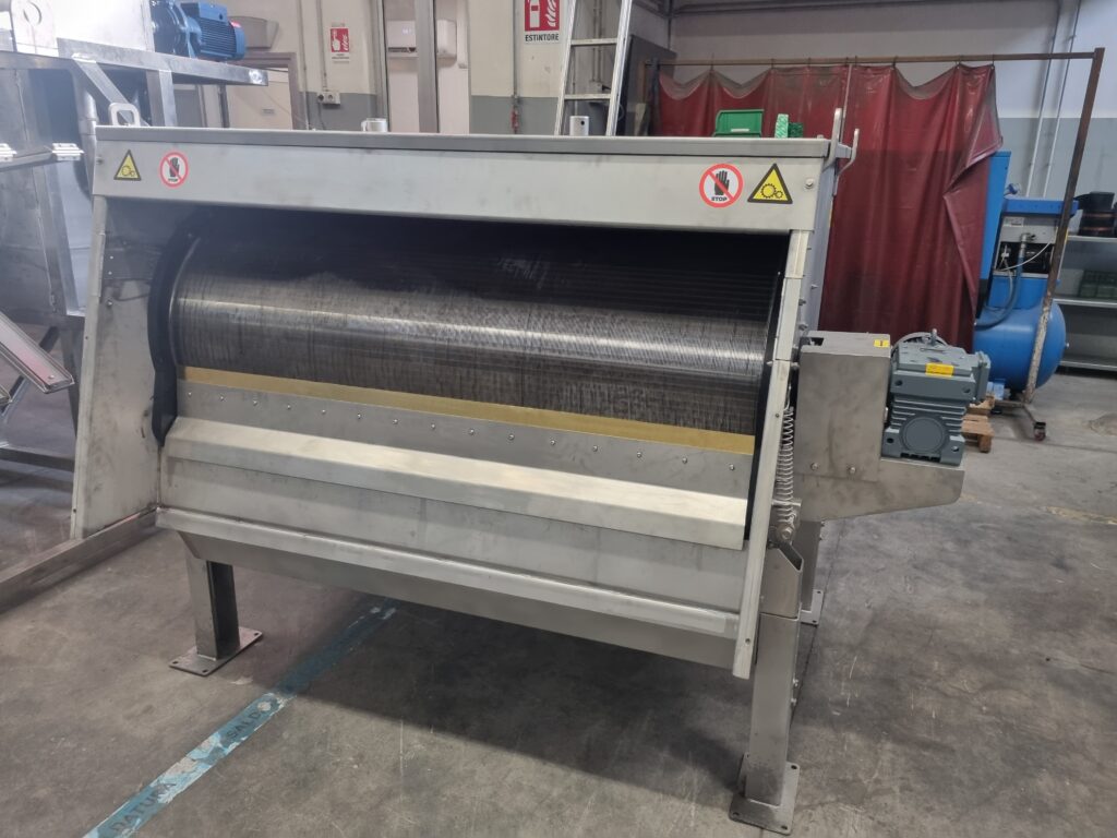 A large industrial stainless steel rotary drum screen machine is standing in a factory setting, surrounded by machinery and safety signs, with a protective cover partially open.