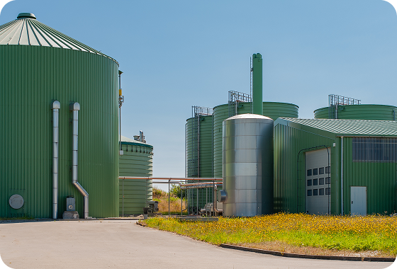 Several large, green industrial tanks and silos stand next to a green metal building, surrounded by wildflowers and grass under a clear blue sky.