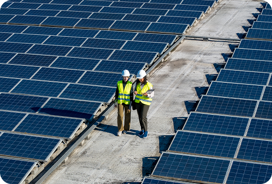 Two people wearing safety vests and hard hats walk between rows of solar panels on a rooftop, inspecting the solar power installation on a sunny day.