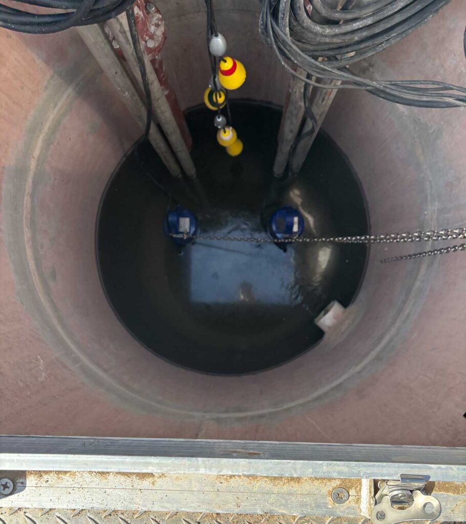 View looking down into a circular well or pit containing water, cables, pipes, two blue submersible pumps, and floating sensor switches. The image is taken from above, showing the interior and equipment setup.